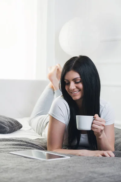 Relajarse en la cama. Hermosa mujer disfrutando de una taza de café y usando su tableta —  Fotos de Stock