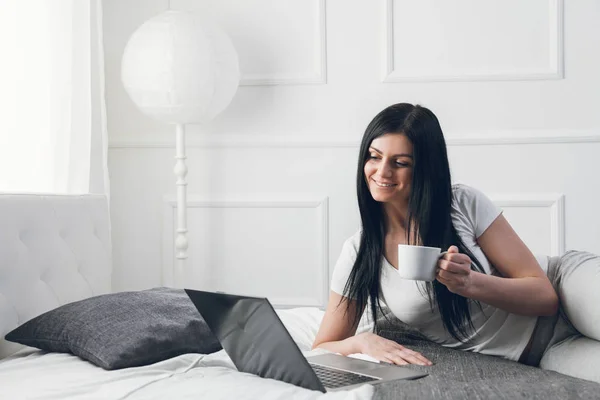 Détente sur le lit. Belle femme profitant d'une tasse de café et utilisant son ordinateur portable — Photo