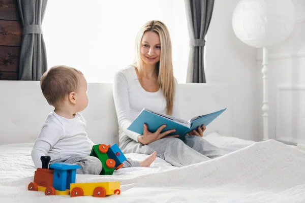 Éducation des enfants. Mère heureuse avec son tout-petit assis sur le lit et lisant un livre — Photo