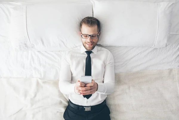 Ansicht von oben. schöner Geschäftsmann mit Brille auf dem Bett liegend SMS von seinem Smartphone — Stockfoto