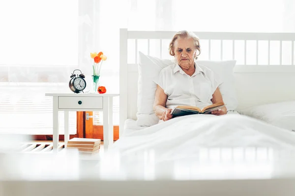 Donna anziana seduta comodamente a letto a leggere il suo libro preferito — Foto Stock