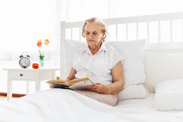 老年妇女舒舒服服地坐在床上读她最喜欢的书 — 图库照片