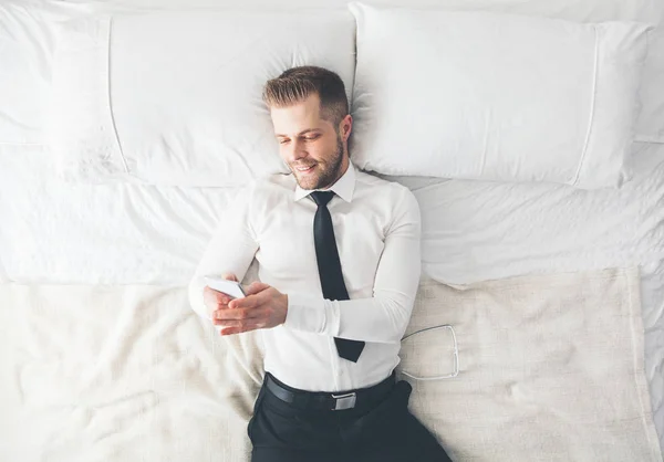 Ansicht von oben. schöner Geschäftsmann, der auf dem Bett liegt und von seinem Smartphone SMS schreibt — Stockfoto