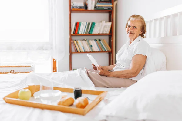 Mulher idosa tomando café da manhã na cama enquanto estiver usando seu tablet — Fotografia de Stock