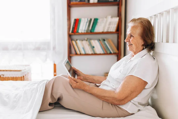 老年妇女舒舒服服地坐在床上，用她的平板电脑 — 图库照片