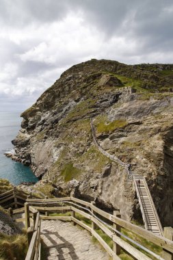 Cornwall, UK: April 14, 2016: Tourists crossing the wooden bridge to Tintagel Island in a vertical format. Tintagel is maintained by English Heritage who are also responsible for other sites across England. clipart