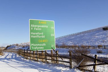 Kışın karlı koşullarla ve mavi gökyüzü arka plan Abergavenny, Hereford ve Monmouth için yön ve mil olarak uzaklık bilgilerle A465 yol işareti.