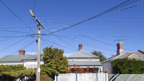 A row of traditional detached houses in a St Kilda suburb with Australian style clapboard walls, fences and verandahs. A telegraph pole has many crossing over cables running into the houses on the street.