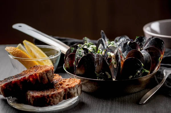 mussels with Dor Blue sauce in pan, bread and lemon slices in bowl on black table