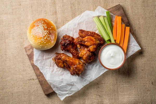 Buffalo chicken wings and bun with sesame on wooden board served with cheese sauce and celery and carrot sticks. top view