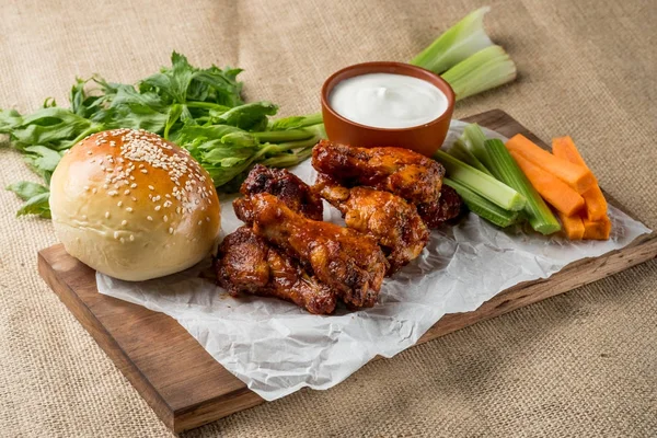 Buffalo chicken wings and bun with sesame on wooden board served with cheese sauce and celery and carrot sticks. top view