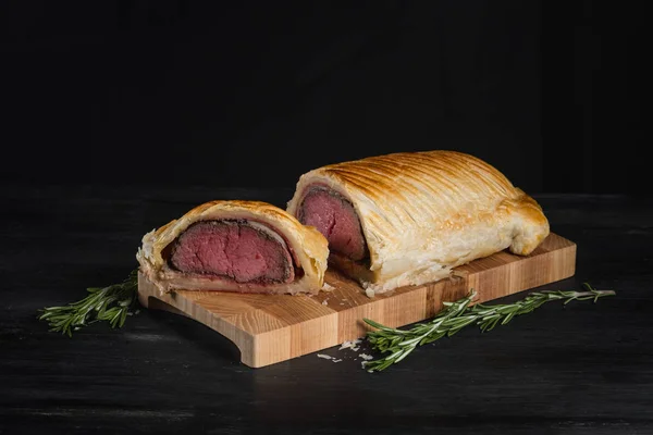Beef wellington with rosemary and spices on wooden board with copy space on black background