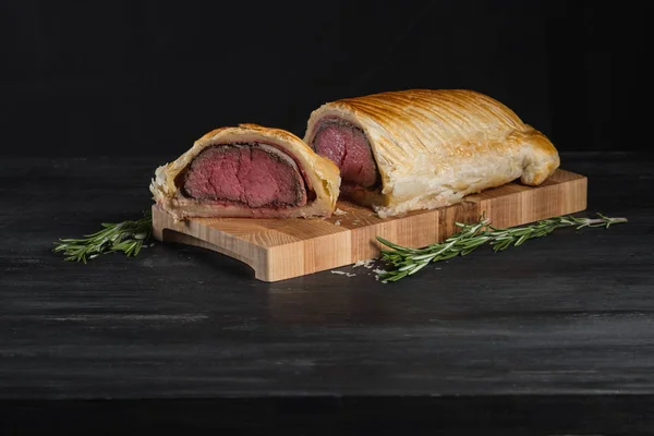 Beef wellington with rosemary and spices on wooden board with copy space on black background