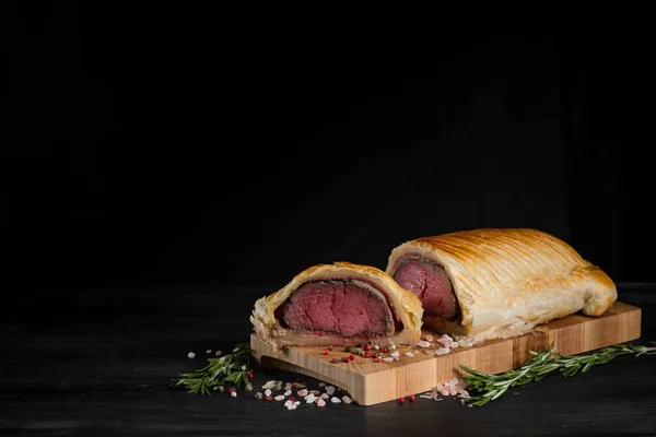 Beef wellington with rosemary and spices on wooden board with copy space on black background, close up view
