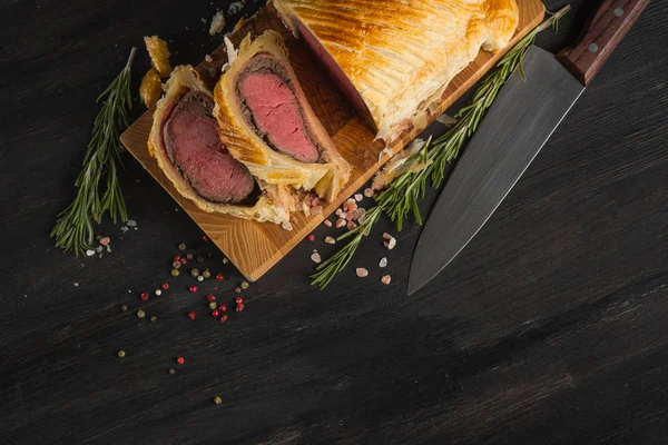 Beef wellington on wooden board with knife, top view with copy space on black background