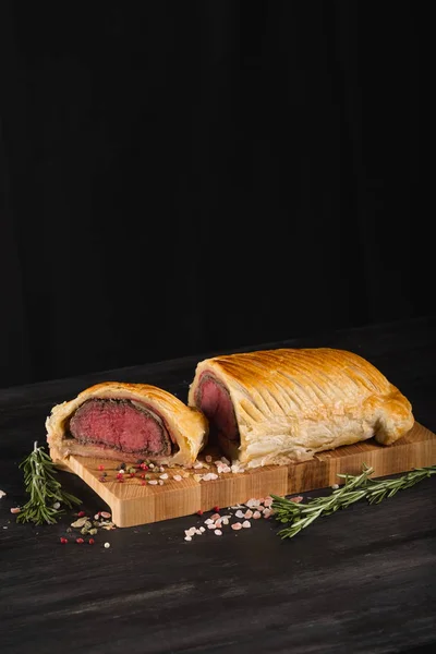 Beef wellington on wooden board with spices and copy space on black background