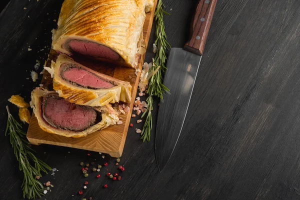 Beef wellington on wooden board with knife, top view with copy space on black background