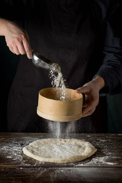 フルイを使って生の生地に小麦粉の粉を注ぐシェフの手のクローズ アップ — ストック写真