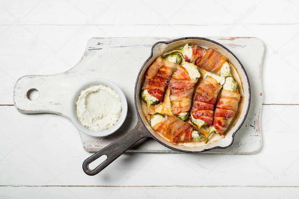 pan with baked peppers stuffed with cheese and bacon on white cutting board with sauce in bowl on wooden background, top view