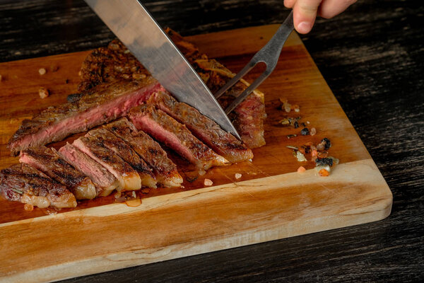 male hands holding knife and carving grilled meat with spices on wooden board background   