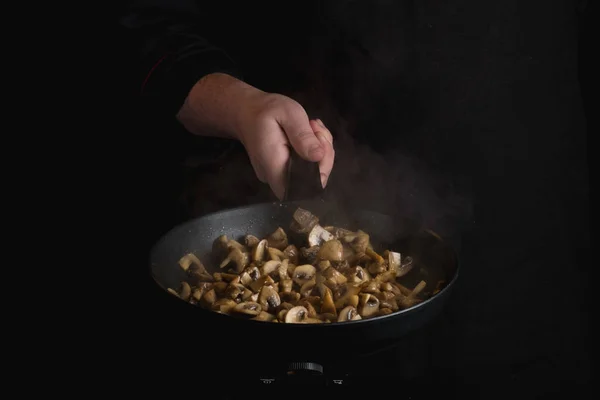 close-up of male chef hand holding black metal frying pan cooking mushrooms on black background