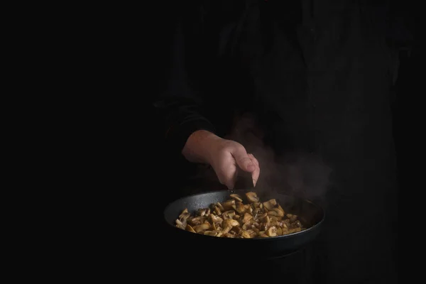 close-up of male chef hand holding black metal frying pan cooking mushrooms on black background