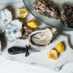 Fresh raw oysters with lemon slices on wooden board with knife and bowl on white table