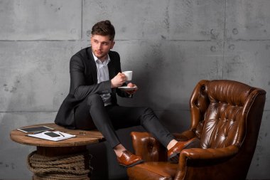 Modern ofis ahşap masada oturan ve kahve fincanı, tam uzunlukta tutan takım elbiseli genç yakışıklı işadamı