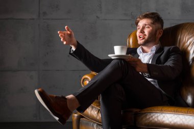 Kahve fincanı tutan deri koltukta oturan takım elbiseli genç işadamı