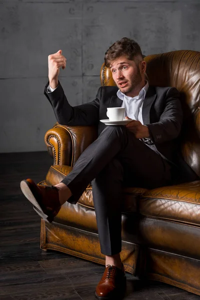 Young businessman in suit sitting on leather sofa holding coffee cup, full length