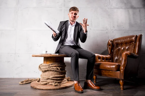 Young angry businessman in suit  sitting on wooden table in modern office holding documents and shouting