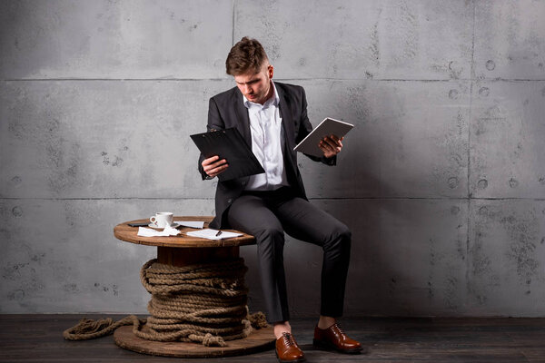 Молодой бизнесмен в костюме сидит на деревянном столе в офисе с цифровыми планшетами и документами
