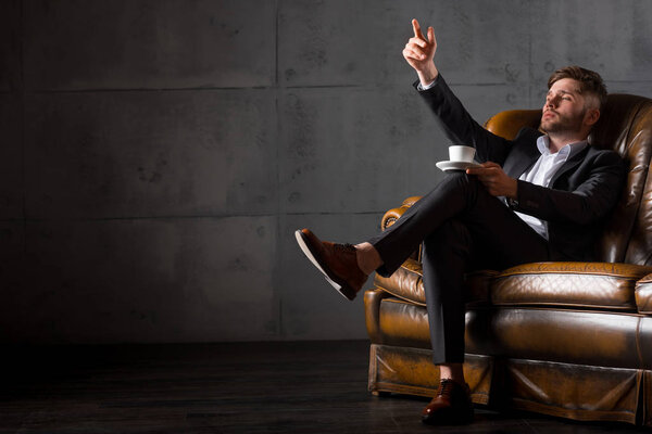 Young businessman in suit sitting on leather sofa holding coffee cup, full length
