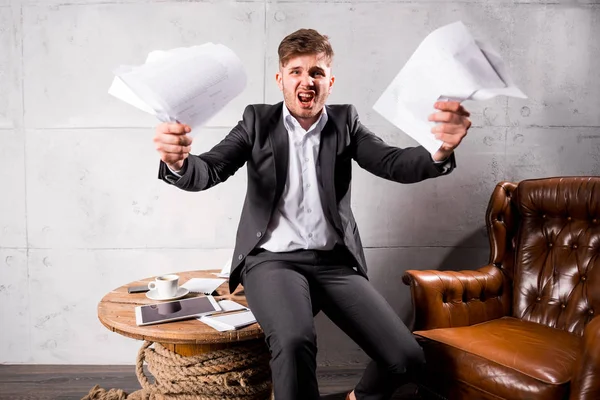 Young angry businessman sitting on wooden table holding documents in hands