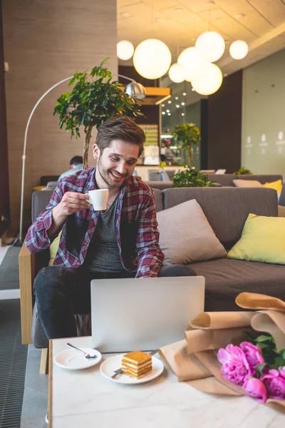 portrait of young bearded man in plaid shirt drink coffee and using laptop  indoor on grey sofa background