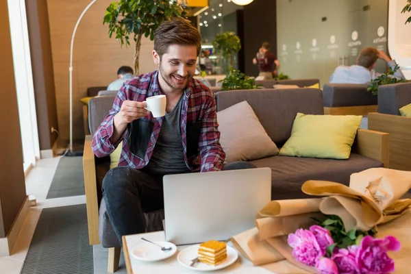 portrait of young bearded man in plaid shirt drink coffee and using laptop  indoor on grey sofa background