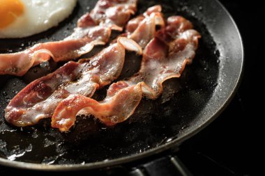 Tavada yumurta ve pastırma pişiyor. Lezzetli geleneksel Amerikan kahvaltısı için. Mutfak siyah arka planda.