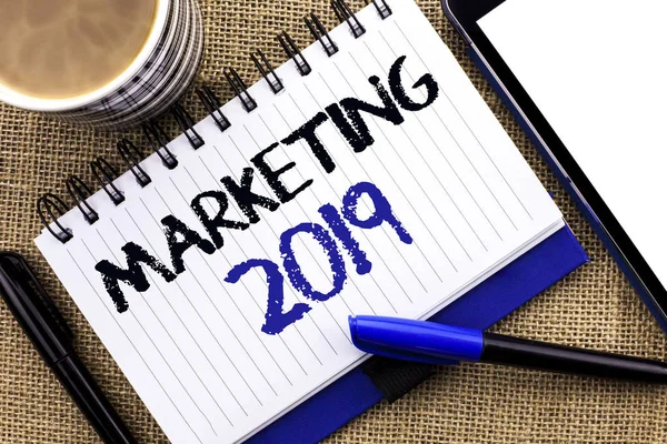 Signo de texto que muestra Marketing 2019. Foto conceptual Mercado de Año Nuevo Estrategias Nuevo comienzo Publicidad Ideas escritas en Notebook Libro sobre el fondo del yute Tablet Coffee Cup and Pens next to it — Foto de Stock