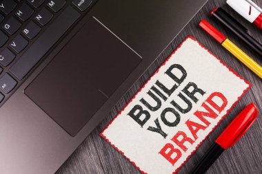 El yazısı metin Build Your marka. Kendi logo sloganı Model reklam E beyaz yapışkan not kağıdına ahşap üzerinde yazılı pazarlama oluşturmak anlam kavramı Laptop kalemler yanında arka plan.