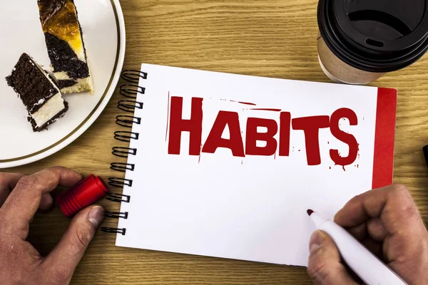 Conceptual hand writing showing Habits. Business photo showcasing Regular tendency or practice Routine Usual Manners Behavior Pattern written by Man on Notebook on wooden background Cake and Cup.