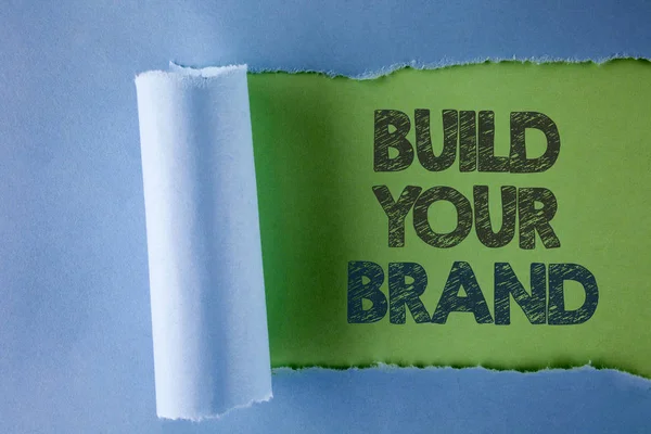 개념적 손을 당신의 브랜드 구축을 보여주는 작성입니다. 비즈니스 사진 텍스트 만들 모델 광고 E 마케팅 일반 녹색 배경에 찢 어 접혀 종이 아래에 서 면 자신의 로고 슬로건. — 스톡 사진