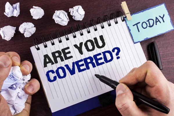 Text sign showing Are You Covered Question. Conceptual photo asking about you insurance health statement written by Man on Notepad holding Marker on wooden background Crumpled Paper Balls