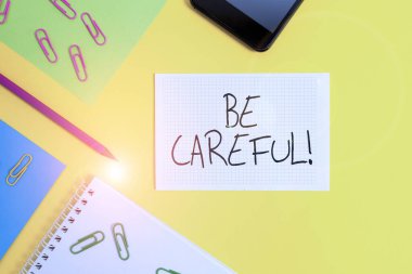 Dikkatli Olun 'u gösteren not yazıyorum. Potansiyel tehlike veya zarardan kaçınmak için iş fotoğrafı gösterimi Kağıt kağıtların kare kalem tokaları akıllı telefon renkli arka plan.