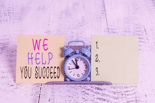 Writing note showing We Help You Succeed. Business photo showcasing Aided Supported Funded someone to reach his dreams Mini blue alarm clock standing above buffer wire between two paper.
