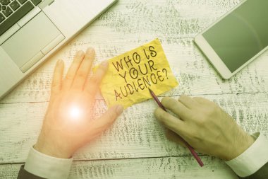 Text sign showing Who Is Your Audience Question. Conceptual photo who is watching or listening to it Hand hold note paper near writing equipment and modern smartphone device. clipart