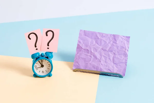 Mini reloj despertador de pequeño tamaño junto a una hoja de papel colocada inclinada. Notepaper en el borde de la mesa vacía multicolor llano telón de fondo. Manera artística de organizar planos pone fotografía — Foto de Stock