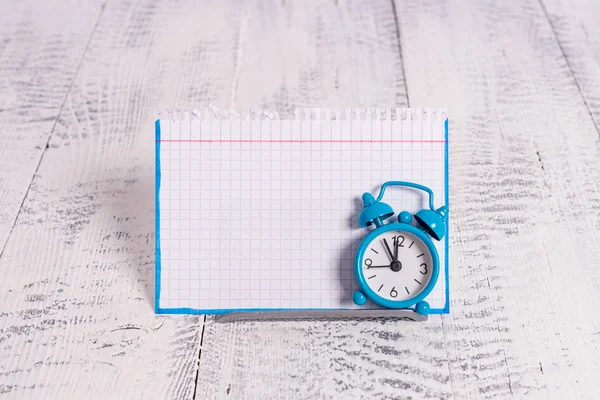 Mini sveglia a colori blu per mostrare il tempo posizionato inclinato di fronte a un notepaper bianco. Classico piccolo orologio in piedi sopra un filo tampone davanti alla carta notazione — Foto Stock