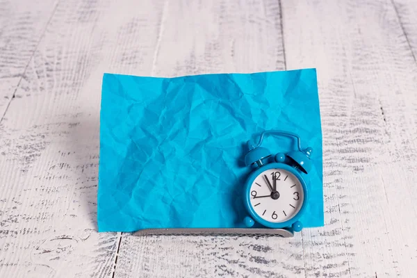 Mini blue colour alarm clock to show time placed tilted in front of one blue notepaper. Classic little watch standing above a buffer wire ahead of notation paper Stock Snímky