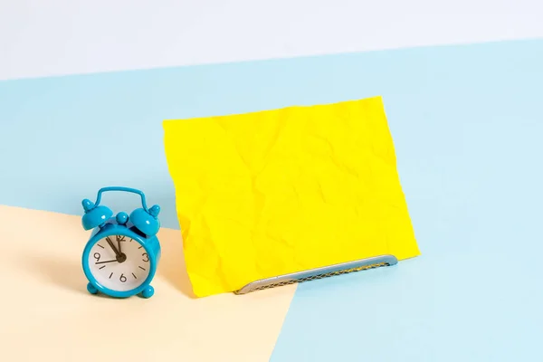 Mini reloj despertador de pequeño tamaño junto a una hoja de papel colocada inclinada. Notepaper en el borde de la mesa vacía multicolor llano telón de fondo. Manera artística de organizar planos pone fotografía — Foto de Stock