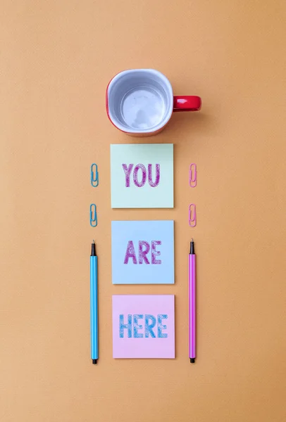 Text sign showing You Are Here. Conceptual photo This is your location reference point global positioning system Coffee cup blank colored sticky note two ballpoints clips yolk background.